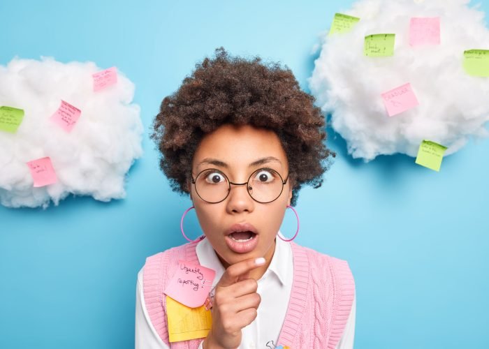 Shocked terrified ethnic female student uses important note uses paper stickers post keeps hand on chin being deeply surprised wears round spectacles formal clothes isolated over blue background